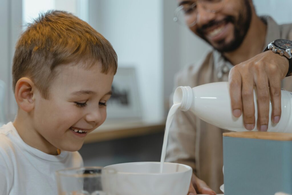 A happy father pouring milk into a bowl for his smiling son during breakfast.